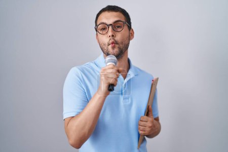 Foto de Hispanic man holding reporter microphone and clipboard making fish face with mouth and squinting eyes, crazy and comical. - Imagen libre de derechos