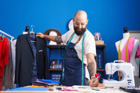 Téléchargez les photos : Jeune homme chauve tailleur souriant confiant tenant veste écriture sur ordinateur portable à l'usine de vêtements - en image libre de droit