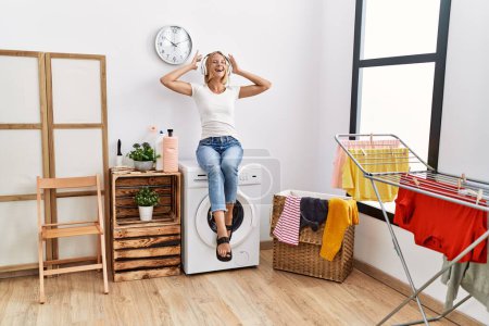 Téléchargez les photos : Jeune femme blonde écoutant de la musique attendant la machine à laver à la buanderie - en image libre de droit