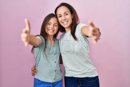 Téléchargez les photos : Jeune mère et fille debout sur fond rose regardant la caméra souriant à bras ouverts pour se faire câliner. expression joyeuse embrassant le bonheur. - en image libre de droit