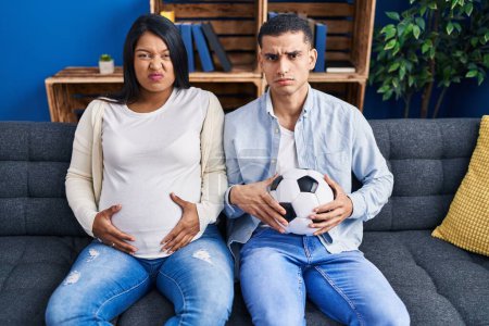 Photo for Young hispanic couple expecting a baby sitting on the sofa holding ball and tummy skeptic and nervous, frowning upset because of problem. negative person. - Royalty Free Image