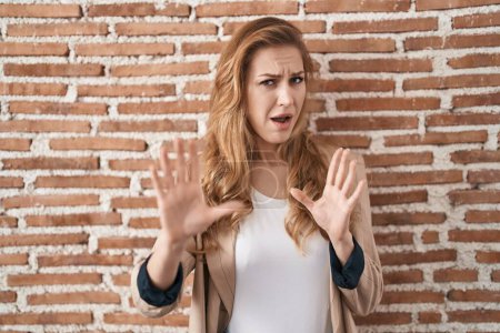 Foto de Beautiful blonde woman standing over bricks wall afraid and terrified with fear expression stop gesture with hands, shouting in shock. panic concept. - Imagen libre de derechos