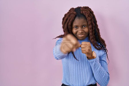 Photo for African woman standing over pink background punching fist to fight, aggressive and angry attack, threat and violence - Royalty Free Image