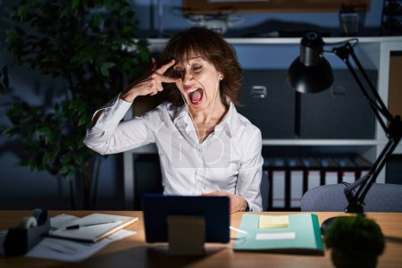 Téléchargez les photos : Middle age woman working at the office at night doing peace symbol with fingers over face, smiling cheerful showing victory - en image libre de droit