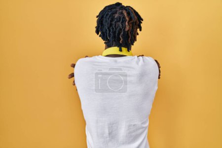Foto de Young african man with dreadlocks standing over yellow background hugging oneself happy and positive from backwards. self love and self care - Imagen libre de derechos