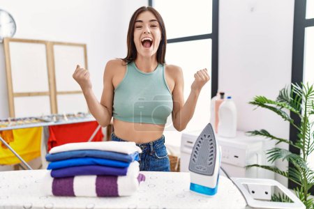 Photo for Young hispanic woman ironing clothes at laundry room celebrating surprised and amazed for success with arms raised and open eyes. winner concept. - Royalty Free Image