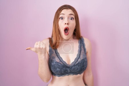Photo for Redhead woman wearing lingerie over pink background surprised pointing with hand finger to the side, open mouth amazed expression. - Royalty Free Image