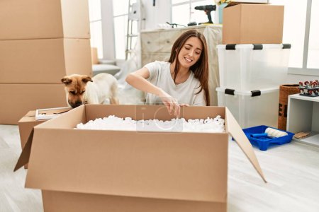 Photo for Young hispanic woman unboxing package sitting on floor with dog at new home - Royalty Free Image