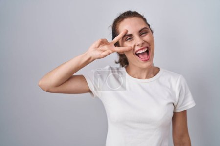 Foto de Beautiful brunette woman standing over isolated background doing peace symbol with fingers over face, smiling cheerful showing victory - Imagen libre de derechos