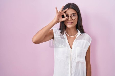 Photo for Brunette young woman standing over pink background wearing glasses doing ok gesture with hand smiling, eye looking through fingers with happy face. - Royalty Free Image