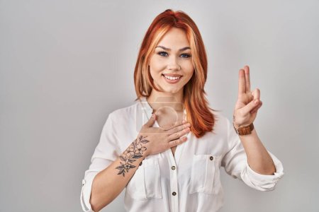 Photo for Young caucasian woman standing over isolated background smiling swearing with hand on chest and fingers up, making a loyalty promise oath - Royalty Free Image