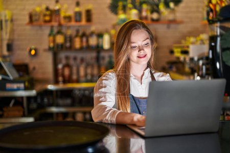 Foto de Mujer joven caucásica camarera sonriendo confiado usando el ordenador portátil en el restaurante - Imagen libre de derechos