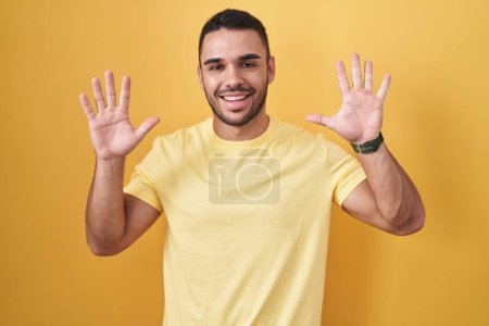 Foto de Young hispanic man standing over yellow background showing and pointing up with fingers number ten while smiling confident and happy. - Imagen libre de derechos