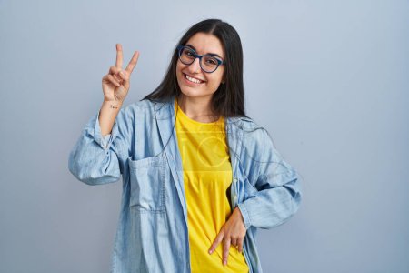 Téléchargez les photos : Jeune femme hispanique debout sur fond bleu souriant regardant la caméra montrant des doigts faisant signe de victoire. numéro deux. - en image libre de droit