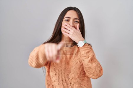 Téléchargez les photos : Young brunette woman standing over white background laughing at you, pointing finger to the camera with hand over mouth, shame expression - en image libre de droit