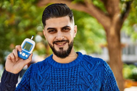 Téléchargez les photos : Jeune homme arabe souriant confiant tenant glucomètre au parc - en image libre de droit
