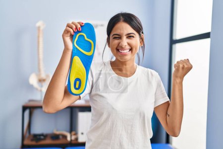 Photo for Young hispanic woman holding shoe insole at physiotherapy clinic screaming proud, celebrating victory and success very excited with raised arm - Royalty Free Image