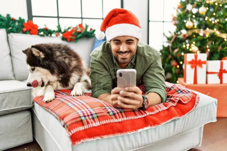 Téléchargez les photos : Young hispanic man using smartphone lying on sofa with dog by christmas tree at home - en image libre de droit
