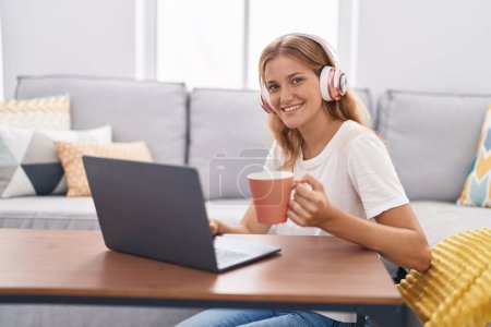 Photo for Young blonde girl watching video on laptop drinking coffee at home - Royalty Free Image