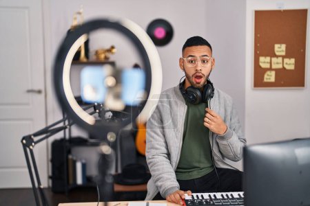 Foto de Young hispanic man playing piano at music studio recording himself scared and amazed with open mouth for surprise, disbelief face - Imagen libre de derechos