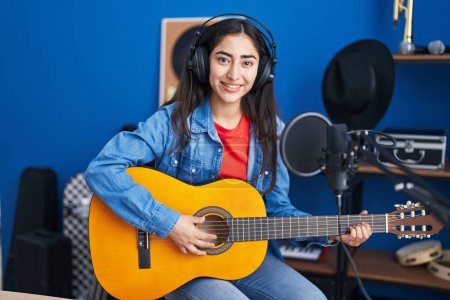 Téléchargez les photos : Young teenager girl playing classic guitar at music studio looking positive and happy standing and smiling with a confident smile showing teeth - en image libre de droit