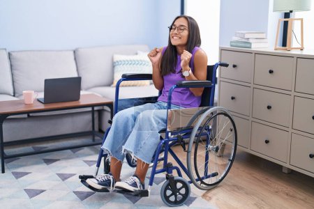 Téléchargez les photos : Young hispanic woman sitting on wheelchair at home excited for success with arms raised and eyes closed celebrating victory smiling. winner concept. - en image libre de droit