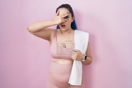 Foto de Young modern girl with blue hair wearing sportswear over pink background peeking in shock covering face and eyes with hand, looking through fingers with embarrassed expression. - Imagen libre de derechos