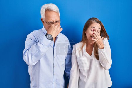 Photo for Middle age hispanic couple standing over blue background smelling something stinky and disgusting, intolerable smell, holding breath with fingers on nose. bad smell - Royalty Free Image
