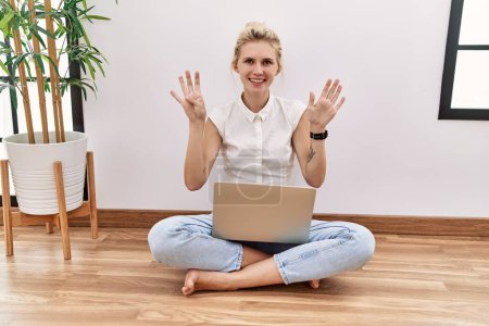 Foto de Young blonde woman using computer laptop sitting on the floor at the living room showing and pointing up with fingers number nine while smiling confident and happy. - Imagen libre de derechos