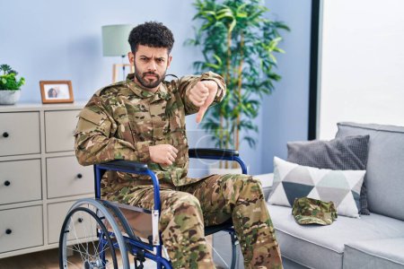 Foto de Arab man wearing camouflage army uniform sitting on wheelchair looking unhappy and angry showing rejection and negative with thumbs down gesture. bad expression. - Imagen libre de derechos