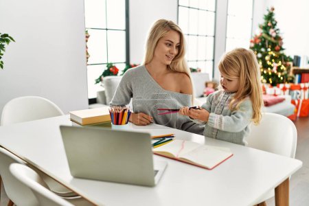Photo for Mother and daughter drawing on notebook using laptop sitting by christmas tree at home - Royalty Free Image