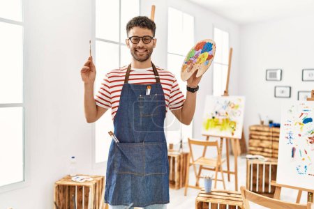 Photo for Young arab artist man smiling happy holding paintbrush and palette at art studio. - Royalty Free Image
