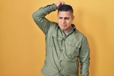 Foto de Hispanic young man standing over yellow background confuse and wondering about question. uncertain with doubt, thinking with hand on head. pensive concept. - Imagen libre de derechos