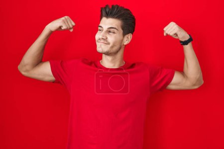 Foto de Joven hombre hispano de pie sobre fondo rojo mostrando los músculos de los brazos sonriendo orgulloso. concepto de fitness. - Imagen libre de derechos