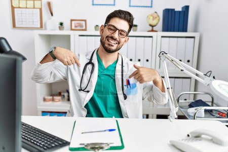 Photo for Young man with beard wearing doctor uniform and stethoscope at the clinic looking confident with smile on face, pointing oneself with fingers proud and happy. - Royalty Free Image