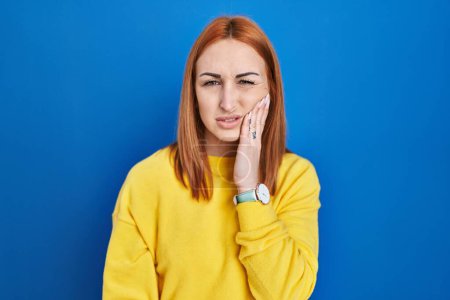 Foto de Young woman standing over blue background touching mouth with hand with painful expression because of toothache or dental illness on teeth. dentist - Imagen libre de derechos