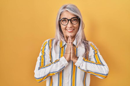 Téléchargez les photos : Middle age woman with grey hair standing over yellow background wearing glasses praying with hands together asking for forgiveness smiling confident. - en image libre de droit