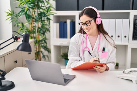 Téléchargez les photos : Young woman wearing doctor uniform listening to music working at clinic - en image libre de droit