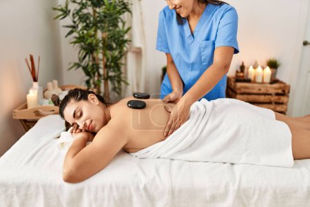 Téléchargez les photos : Deux femmes thérapeute et patient ayant séance de massage du dos en utilisant des pierres noires au centre de beauté - en image libre de droit