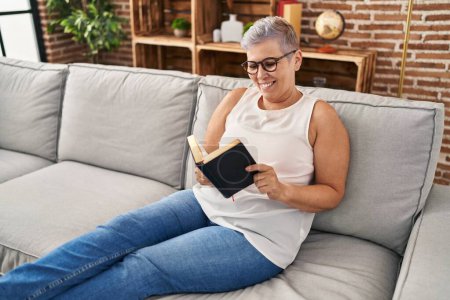 Téléchargez les photos : Femme d'âge moyen lisant la bible assise sur le canapé à la maison - en image libre de droit