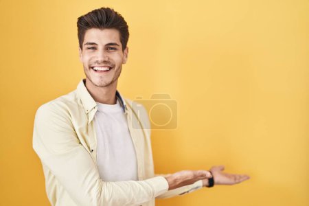 Foto de Joven hispano de pie sobre fondo amarillo invitando a entrar sonriente natural con la mano abierta - Imagen libre de derechos