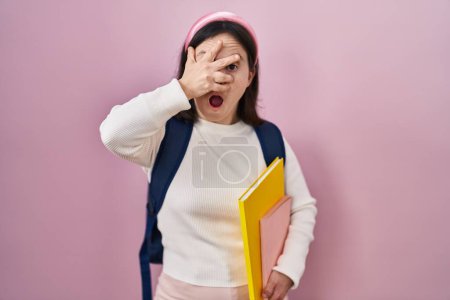 Foto de Woman with down syndrome wearing student backpack and holding books peeking in shock covering face and eyes with hand, looking through fingers with embarrassed expression. - Imagen libre de derechos
