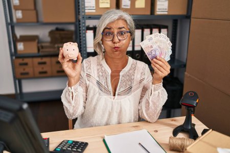 Foto de Middle age woman with grey hair working at small business ecommerce holding piggy bank and zloty puffing cheeks with funny face. mouth inflated with air, catching air. - Imagen libre de derechos