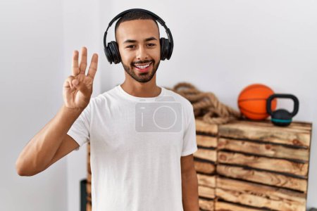 Téléchargez les photos : African american man listening to music using headphones at the gym showing and pointing up with fingers number three while smiling confident and happy. - en image libre de droit