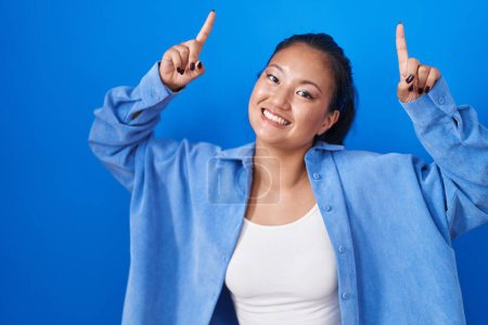 Téléchargez les photos : Asiatique jeune femme debout sur fond bleu sourire étonné et surpris et pointant vers le haut avec les doigts et les bras levés. - en image libre de droit
