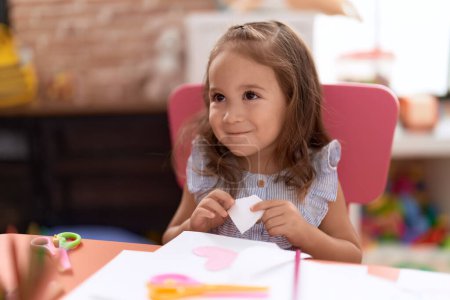 Téléchargez les photos : Adorable hispanic girl student sitting on table cutting paper at kindergarten - en image libre de droit