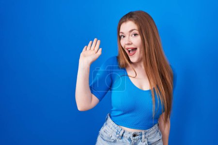 Téléchargez les photos : Redhead woman standing over blue background waiving saying hello happy and smiling, friendly welcome gesture - en image libre de droit
