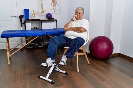 Photo for Senior caucasian man at physiotherapy clinic using pedal exerciser with hand on chin thinking about question, pensive expression. smiling with thoughtful face. doubt concept. - Royalty Free Image