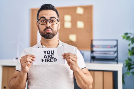 Photo for Young hispanic man with beard holding you are fired banner at the office puffing cheeks with funny face. mouth inflated with air, catching air. - Royalty Free Image