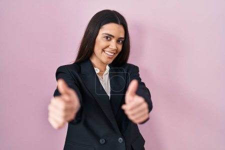 Foto de Young brunette woman wearing business style over pink background approving doing positive gesture with hand, thumbs up smiling and happy for success. winner gesture. - Imagen libre de derechos
